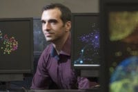 A photo shows a man in purple dress shirt behind computer monitors