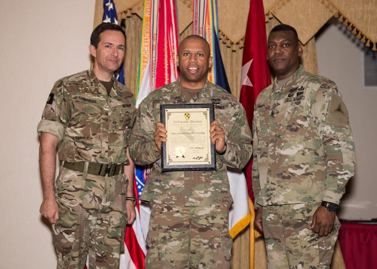 Three male soldiers dressed in camouflage uniforms. One soldier holds certificate.