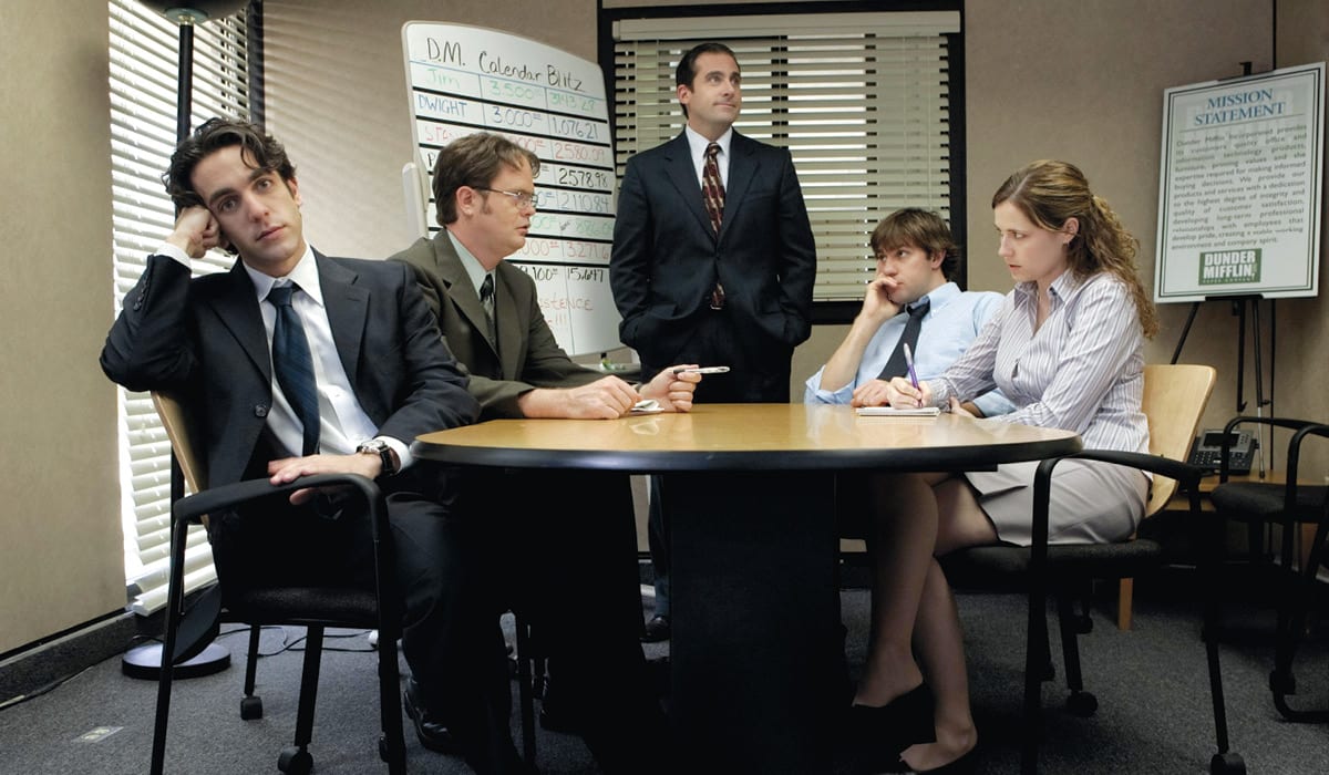 Scene from the TV show The Office showing manager Michael Scott presiding over a meeting with four disinterested employees.
