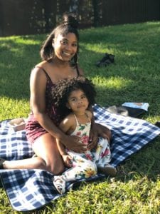 A black woman and her daughter sits on a blue-and-white checkered blanket.