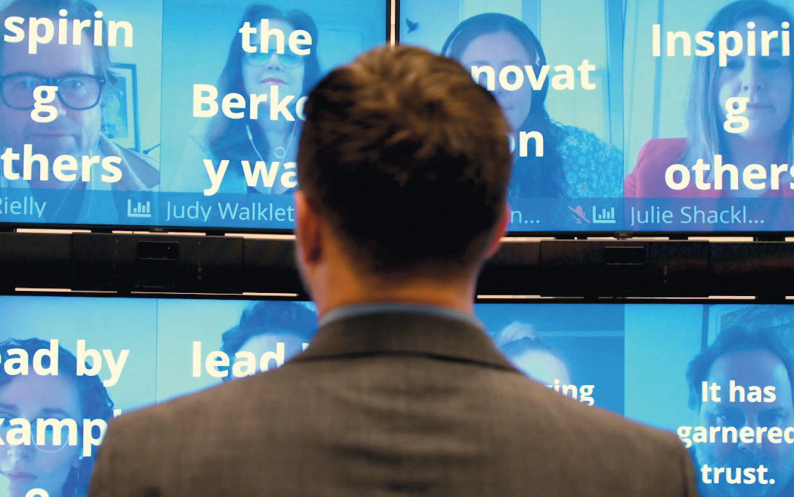 Close-up of the back of a teacher standing in front of a wall of screens showing people overlaid with their answers to a poll. The answers include Inspiring Others and Lead by Example.