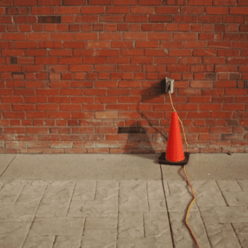 an orange cone near a brick wall. An electric cord plugged into brick wall.