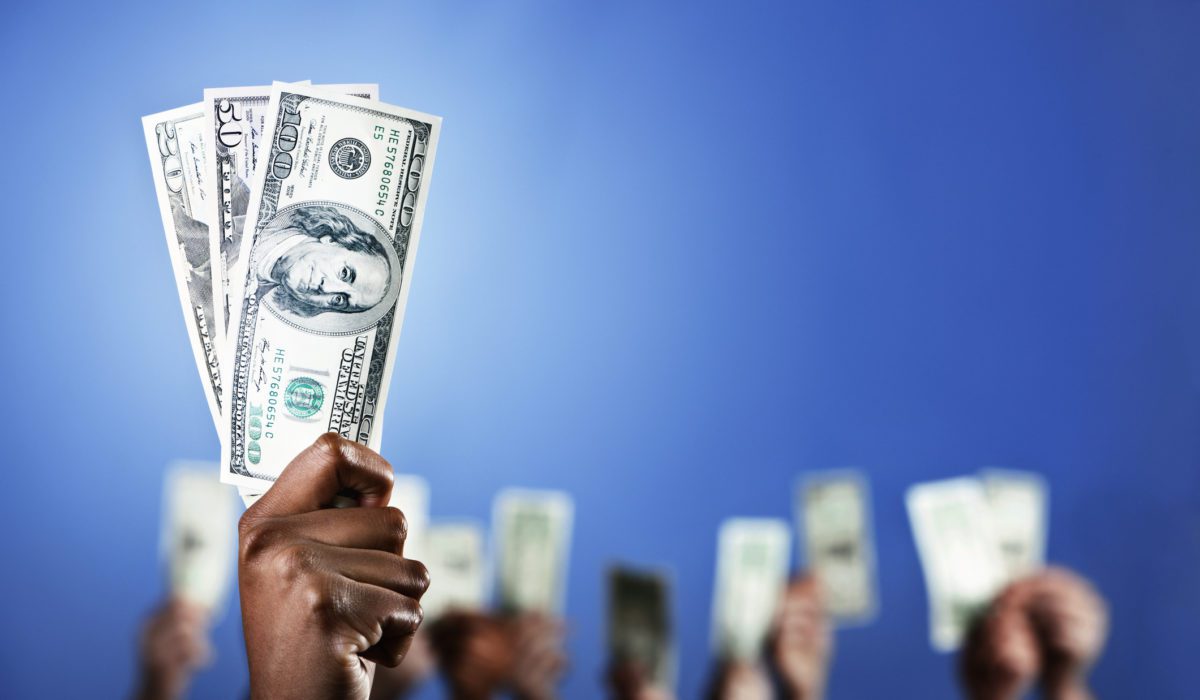 A hand holds up a bundle of dollars while many other hands do the same in the background