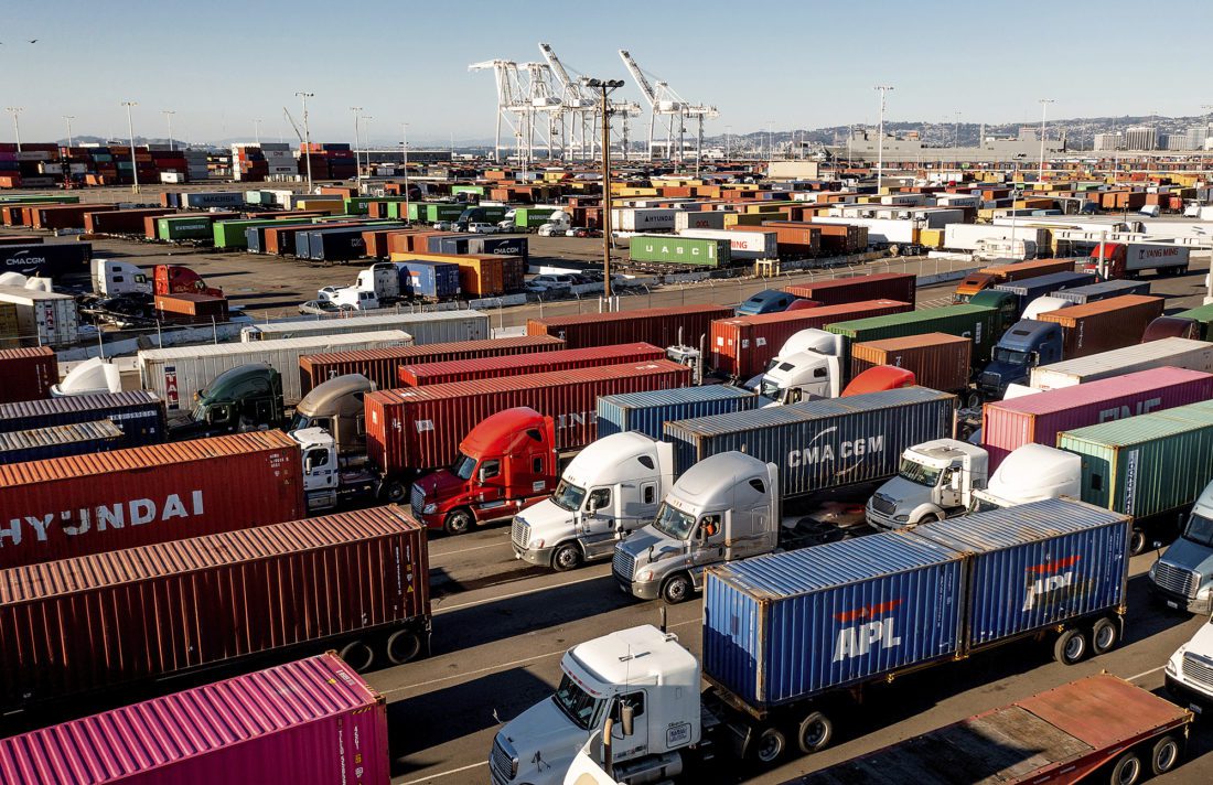 Trucks lined up to enter the Port of Oakland. Shipping containers and cranes are visible in the background.