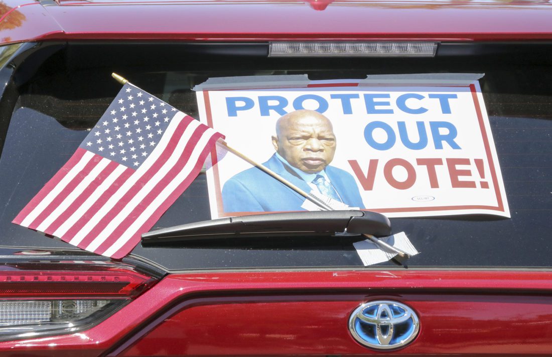 A car with a sign reading "Protect Our Vote!" waits in line at a voting rights demonstration.