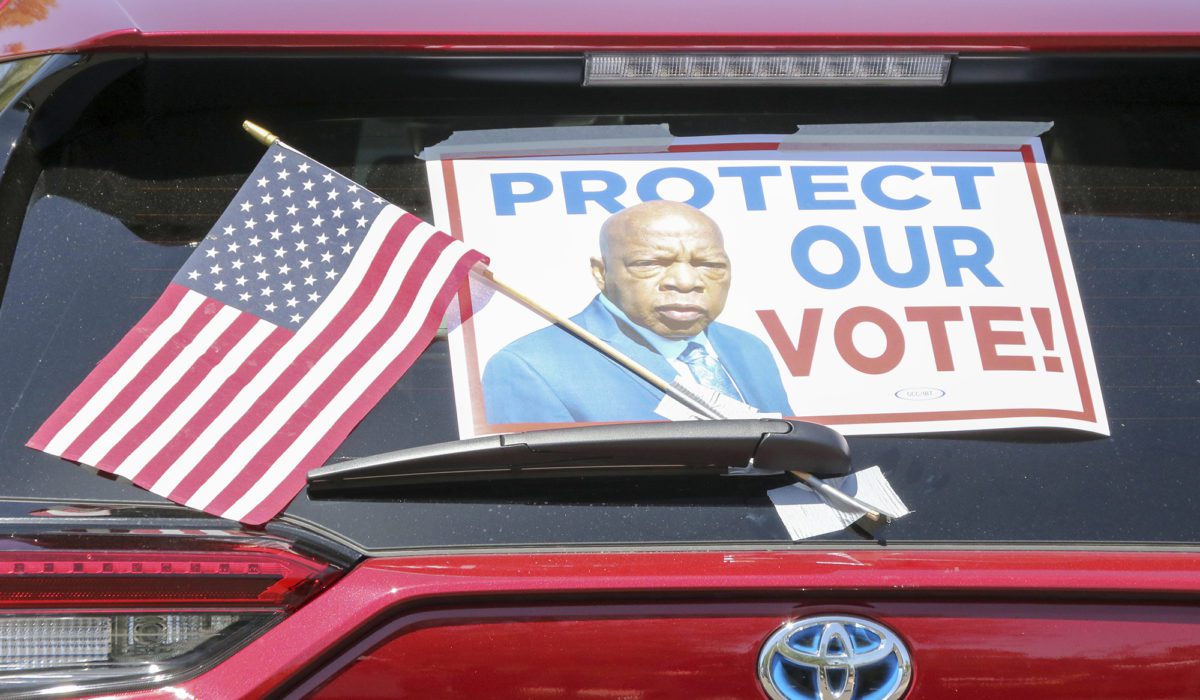 A car with a sign reading "Protect Our Vote!" waits in line at a voting rights demonstration.