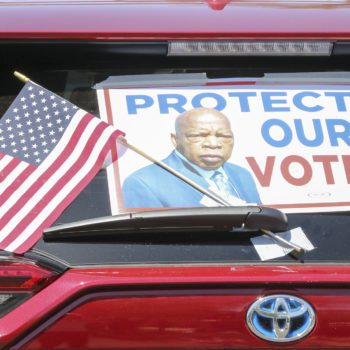 A car with a sign reading "Protect Our Vote!" waits in line at a voting rights demonstration.