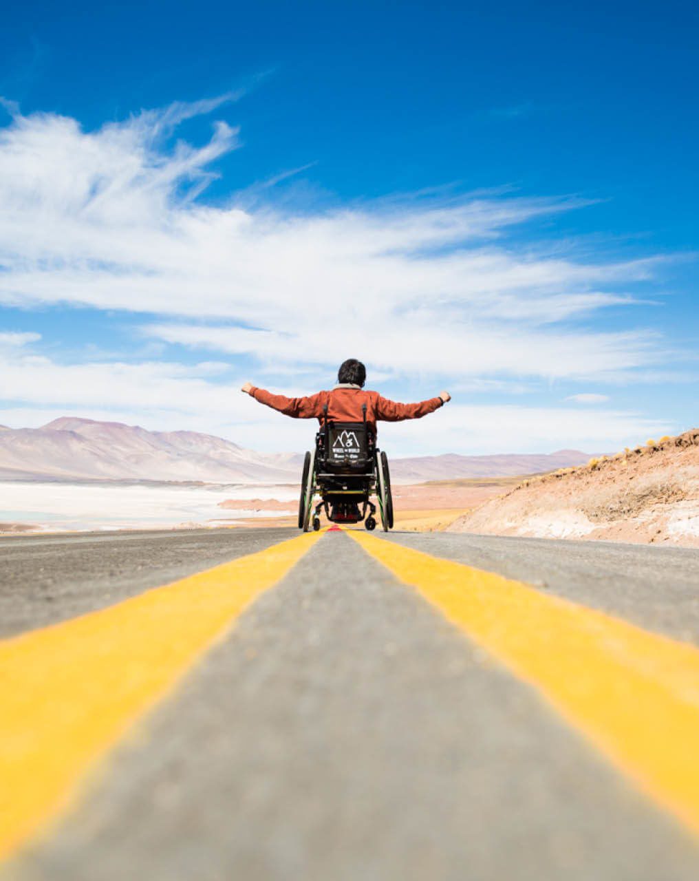 Man in a wheelchair riding along the center line of a road with his arms outstretched.