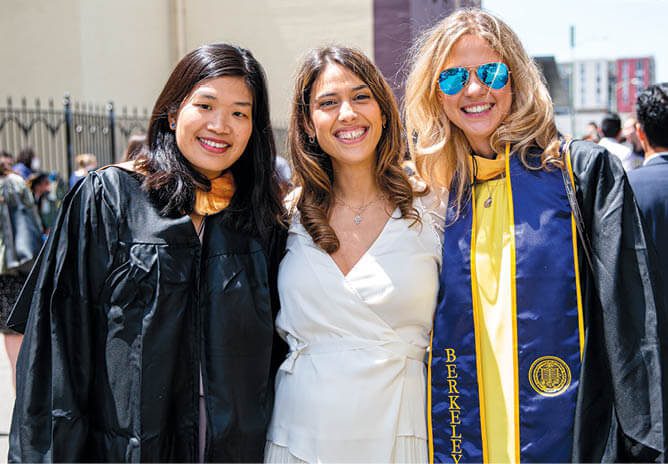 Three women standing together; two are wearing graduation gowns.