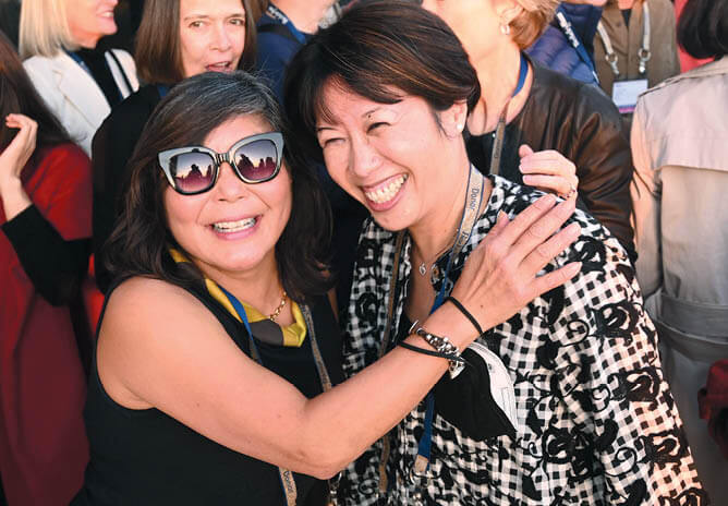 Two women standing together and smiling with a crowd of people behind them.