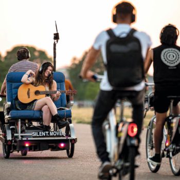 A guitarist being driven on a pedal cart followed by people on bikes wearing headphones.