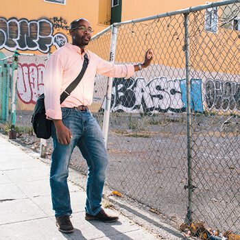 Man with one arm on a chain-link fence and a wall of graffiti behind him.