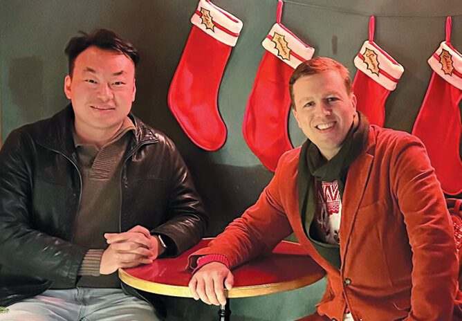 Two men seated in front of a row of Christmas stockings and smiling.