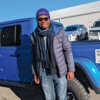 Oseyi Ikuenobe wearing a scarf and hat and standing in front of a blue truck.