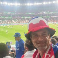Pascal Hoffmann in a bucket hat in the stands of a soccer stadium, with a match happening behind him.