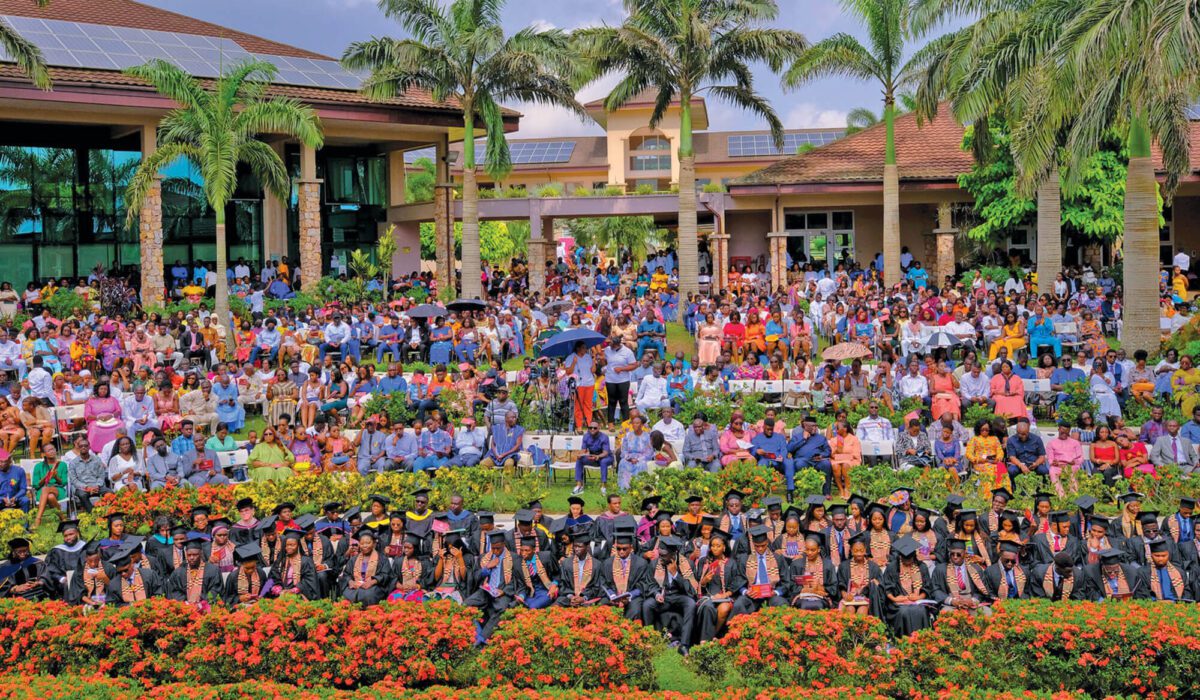 Graduating students in caps and gowns at the front of the picture with rows of audience members behind them as well as university buildings and palm trees.