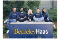 5 Haas Students sitting on Berkeley Haas sign. Smiling.