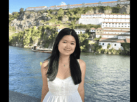 Female Student. Smiling in a white summer dress. Backdrop is water and coastal town.