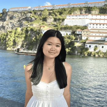 Female Student. Smiling in a white summer dress. Backdrop is water and coastal town.