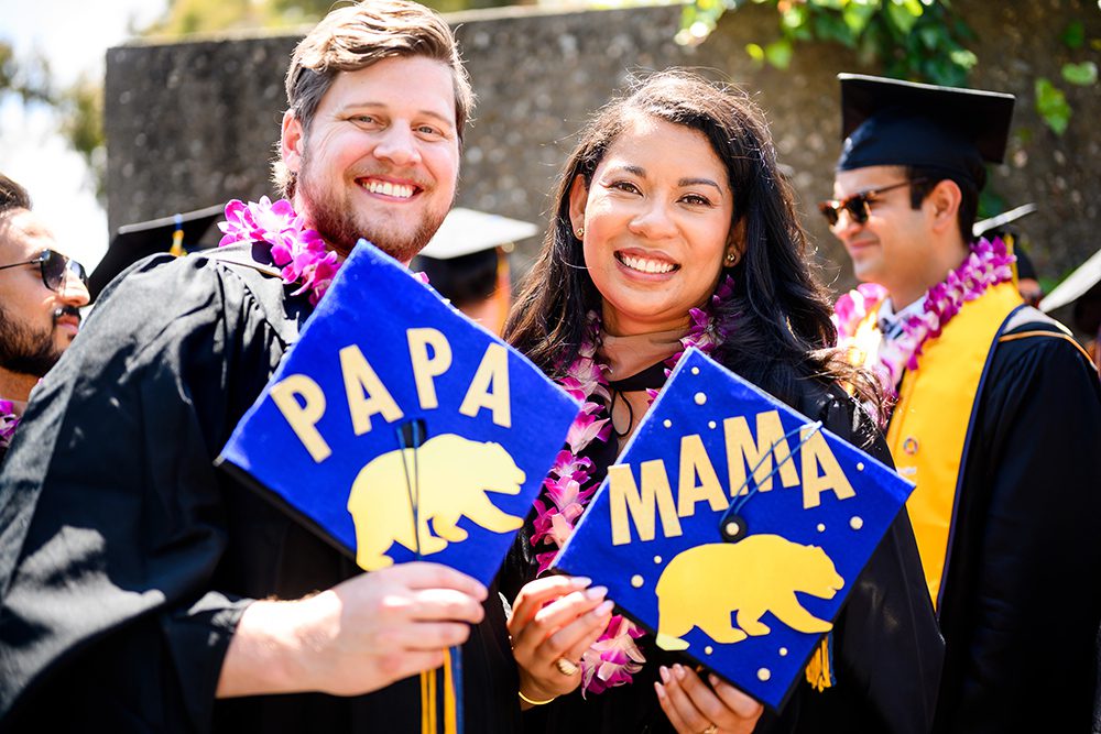 A man holding a graduation cap that says Papa with a picture of a bear standing next to a woman holding a cap that says Mama with a picture of a bear.