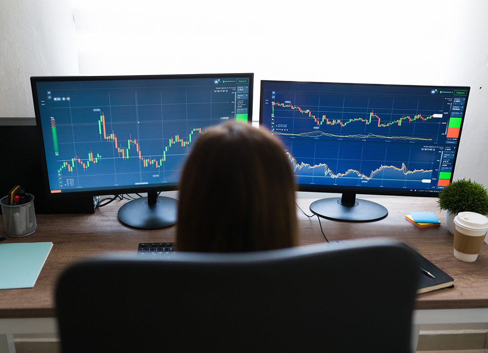 Photo of a professional woman looking at two monitors showing stock prices