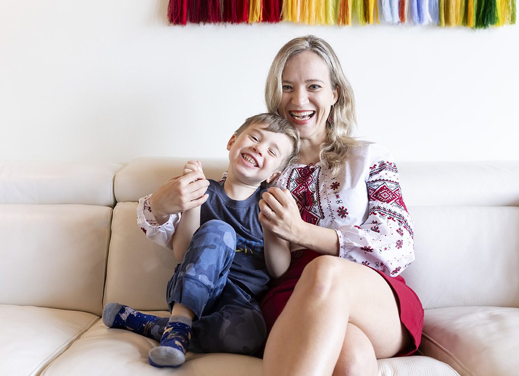 A woman with blond hair sits on a white sofa with her young son. Both are smiling.