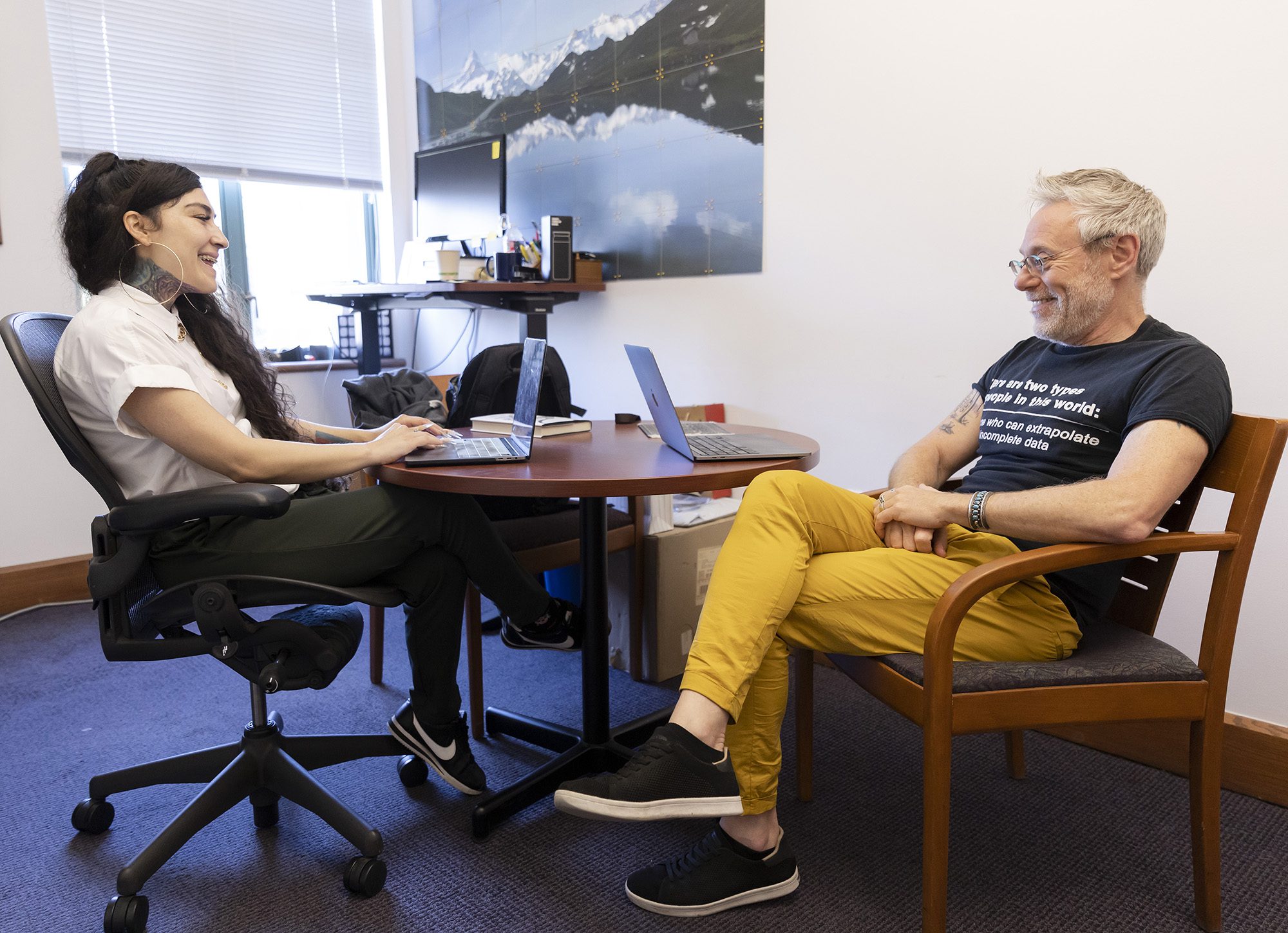 A woman with long dark hair wearing a white blouse sits in an office chair talking to a man wearing black t-shirt and yellow pants