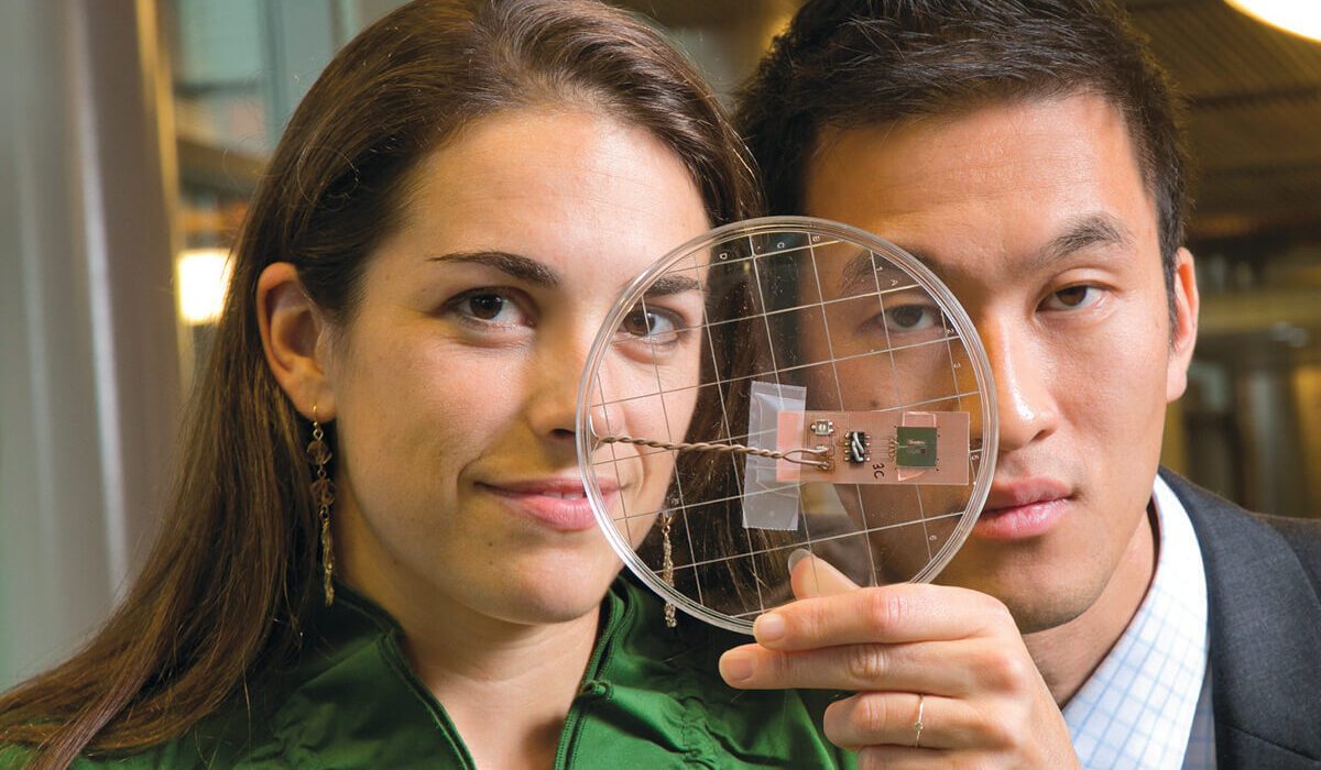 A woman and a man; one is holding up a small electronic device in what looks to be a petri dish.