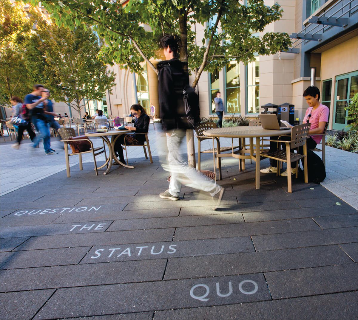 Berkeley Haas courtyard showing Question the Status Quo etched into the pavement. Some students sit at tables others are walking; the photo shows the motion of their bodies.