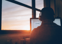 Photo of a man, whose back is facing the camera, wearing headphones and working on a computer at dusk.