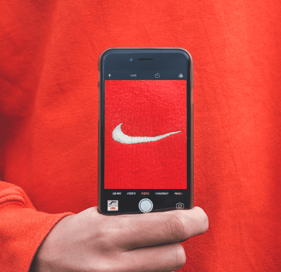 Photo of an arm wearing a red top holding a smart phone with a Nike sign in the middle of the screen against a red backdrop.