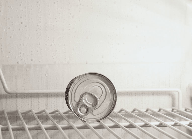 Photo of a one can of food or beverage on its side in an empty refrigerator on an empty refrigerator shelf.