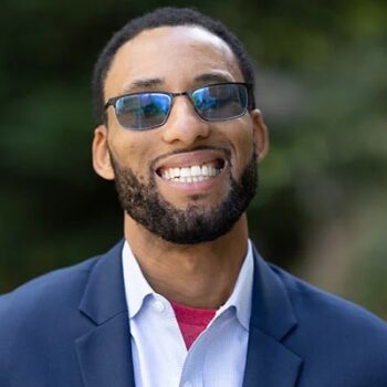 African American male smiling, wearing dark blue blazer, light blue button-up shirt with red tee shirt underneath.