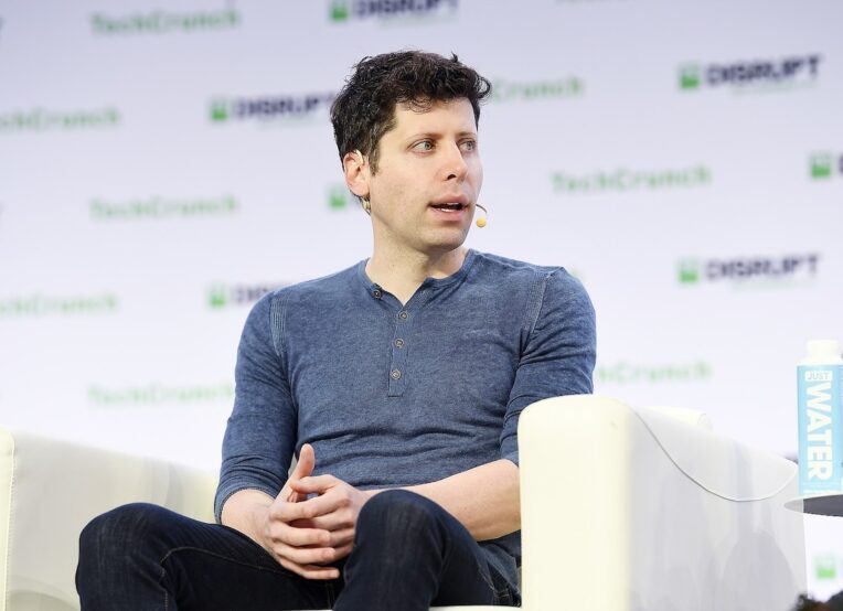 Photo of OpenAI CEO, Sam Altman, at a talk sitting in a white chair wearing a blue shirt and dark pants.