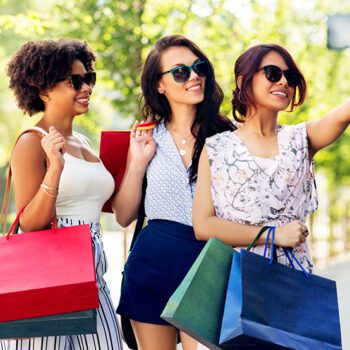 Three women with shopping bags taking a selfie in city.