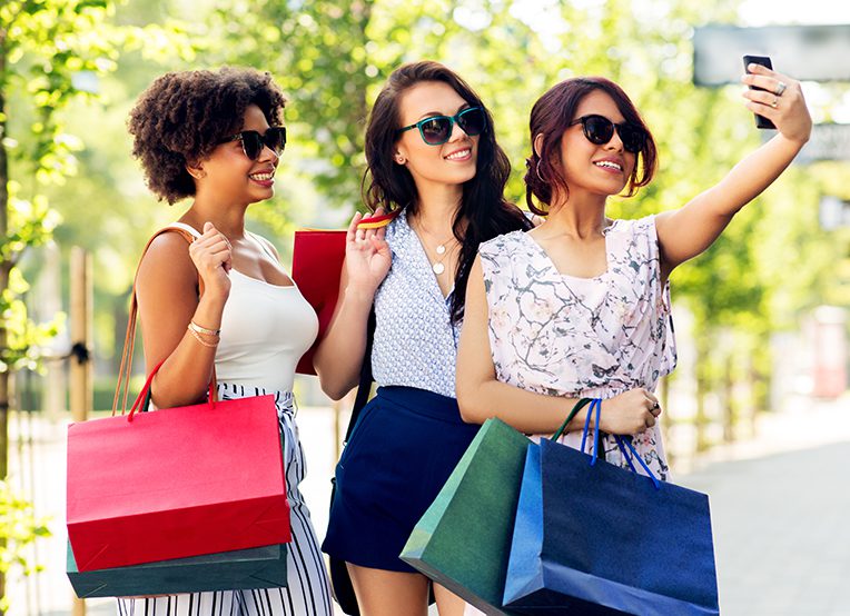 Three women with shopping bags taking a selfie in city.