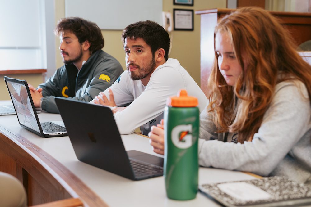 three students sitting with laptops in a classroom