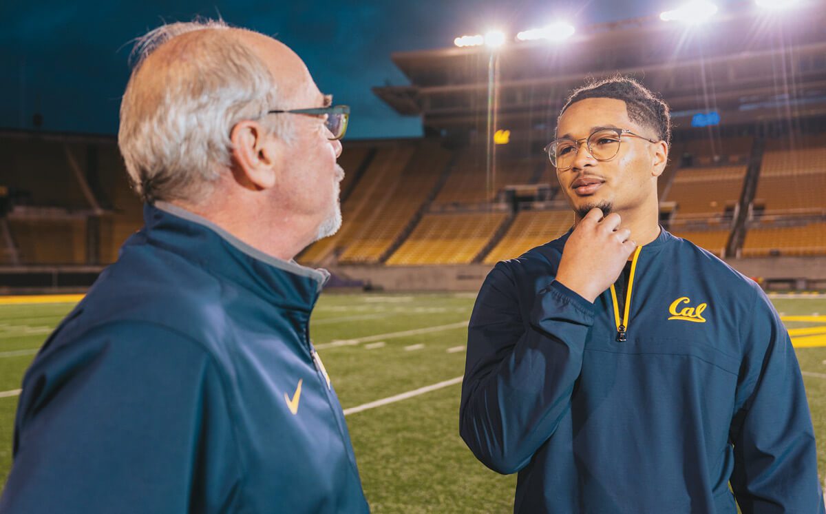 Two men, one older and one younger, both in Cal zip-up shirts, standing on the football field in California Memorial Stadium talking.
