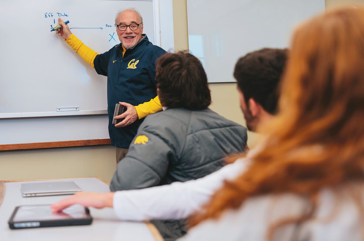 A man in a Cal zip-up shirt standing at a whiteboard, teaching a financial literacy class to student-athletes.