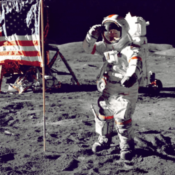 A photo of an Astronaut on the moon standing to the right of a USA flag.