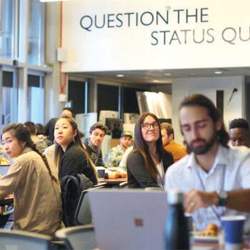 Students sitting at tables in an entrepreneurship class. Above them is a message on the wall reading Question the Status Quo.