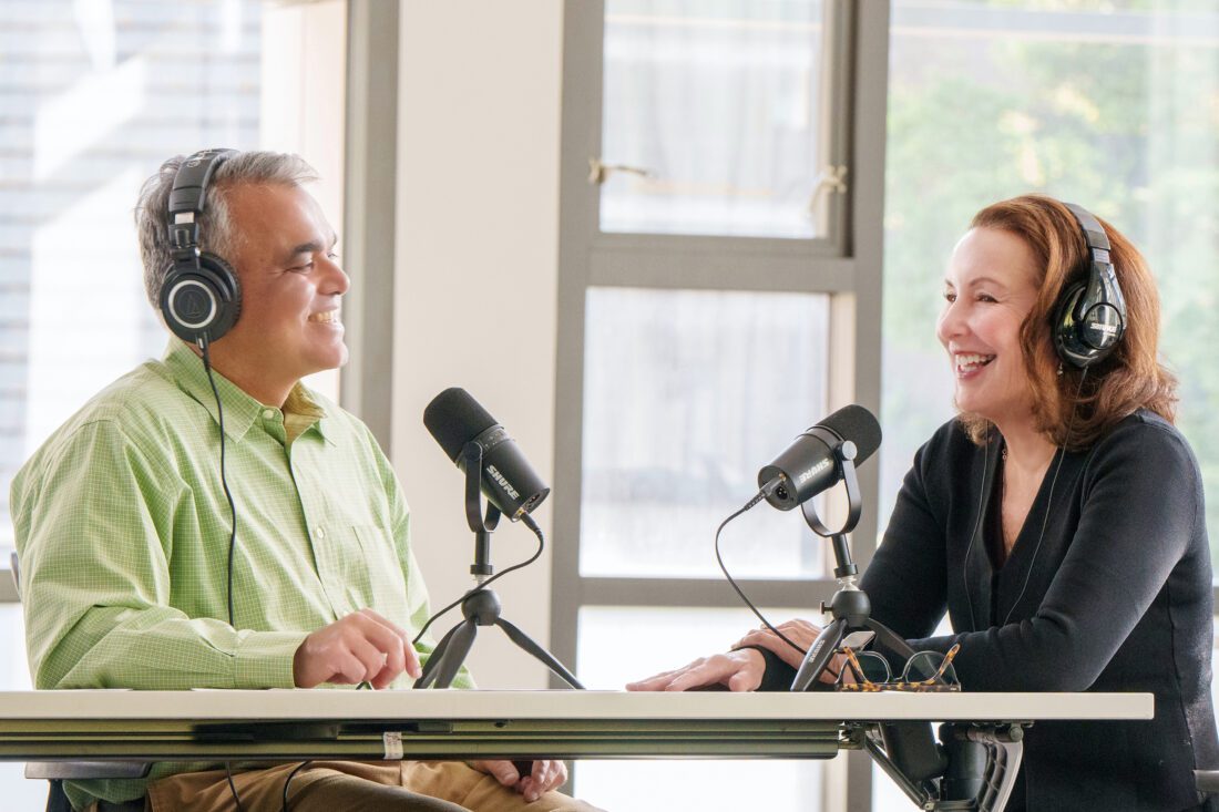 A man and woman sit at a table wearing headphones and speaking into podcasting microphones. 