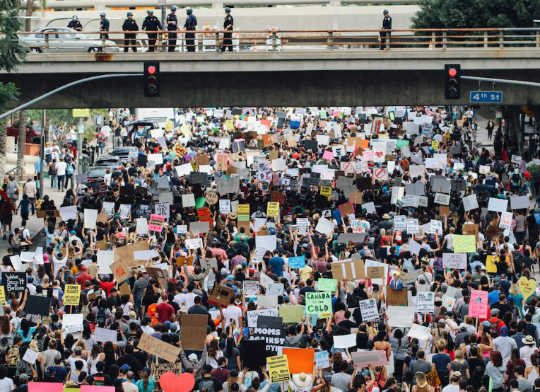 Photo of a large crowd on a street, protesting. Above the protest on a ramp are police officers.
