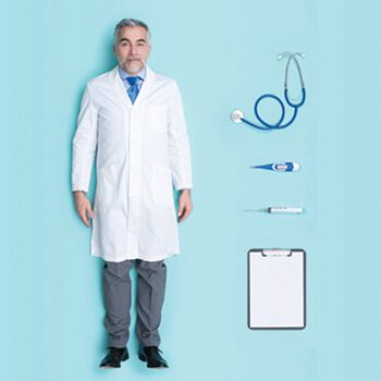 Man dressed as a doctor. Next to him are a stethoscope, thermometer, scope, and clipboard. Credit: istock.