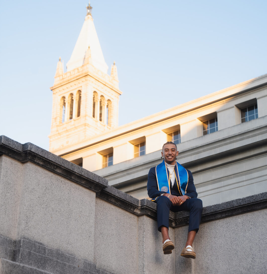 man sitting on a wall wearing commencement sash with campanille in back