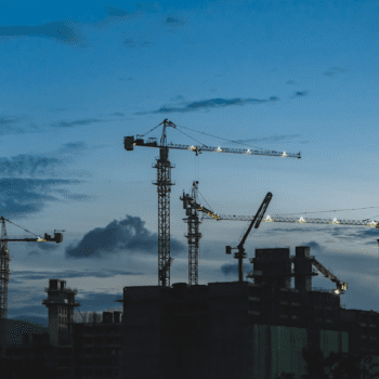 Photo of 3 large cranes towering over a building/pant at dusk.