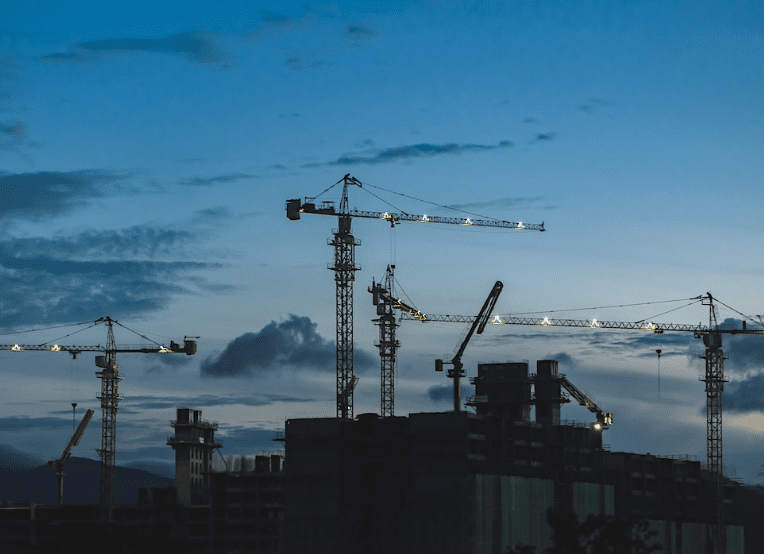 Photo of 3 large cranes towering over a building/pant at dusk.