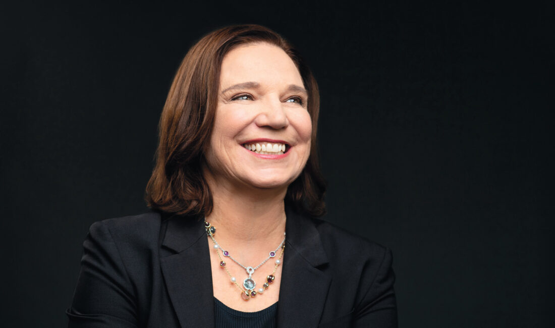 A smiling woman wearing a dark suit and necklaces photographed against a black background.