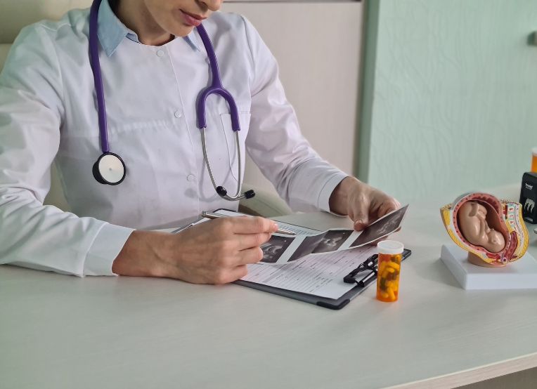 A female doctor with stethoscope around her neck examines a sonogram image of a fetus. A prescription bottle and a model of a uterus is visible on the table.