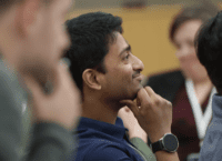 Photo of Students in a classroom. The student in the background is blurred but looks to be a female. The two students in the foreground are male, both looking either at the professor or the board, focused with hand resting on chin.